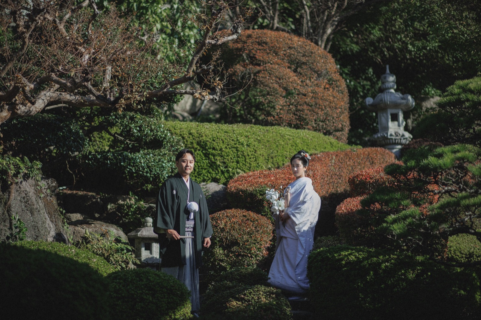日本庭園に佇む、羽織袴と白無垢姿の新郎新婦。石灯籠と緑に囲まれて。