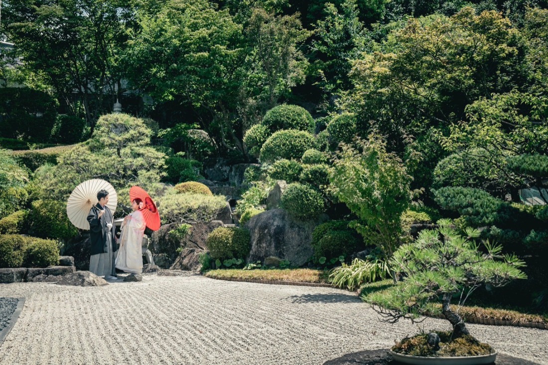 白い傘と赤い傘を手にした新郎新婦。石組みと松のある庭園にて。